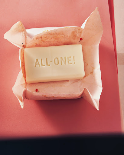 A bar of soap rests in wrinkled paper wrapping on a pink surface. Embossed text on the soap reads "ALL-ONE!"