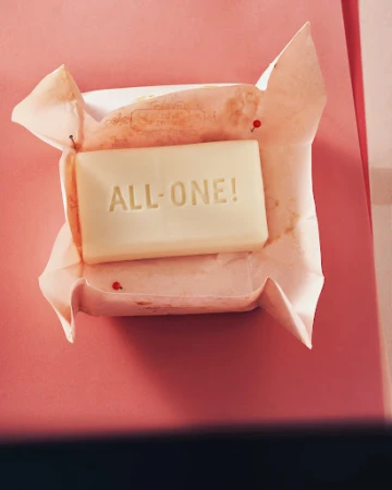 A bar of soap rests in wrinkled paper wrapping on a pink surface. Embossed text on the soap reads "ALL-ONE!"