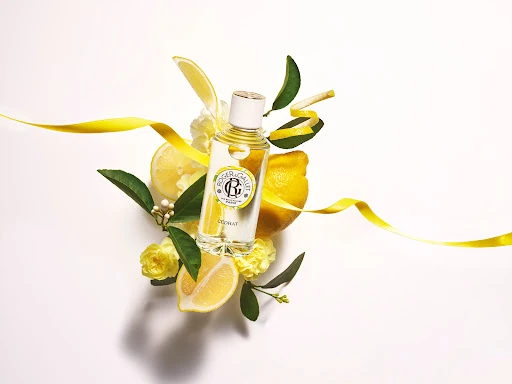 Product shot of Roger & Gallet L'Homme Cedrat perfume bottle surrounded by lemons, lemon peels, flowers, ribbon against white background.