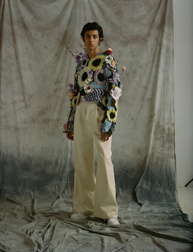 A person posing in wide-leg pants and floral-embellished top against a mottled backdrop, fashion forward, studio lighting, full body shot.