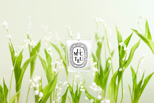 Close-up studio shot of lit Diptyque candle surrounded by lily of the valley flowers, set against a soft, blurred background.