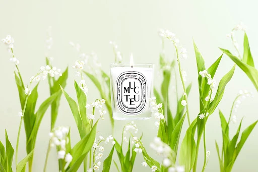 Close-up studio shot of lit Diptyque candle surrounded by lily of the valley flowers, set against a soft, blurred background.