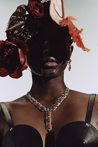 Portrait of a dark-skinned woman with flower headdress and large coral jewelry against a white background in a high-fashion editorial style.