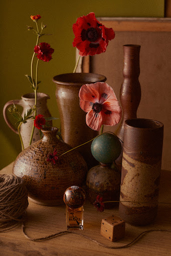 Still life featuring a collection of vases, ceramic vessels, flowers, a glass perfume bottle, and a wooden die on a wooden surface.