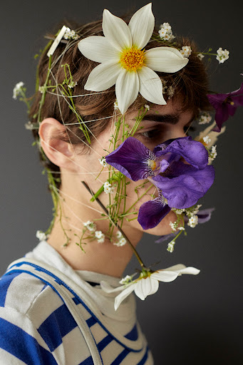 Conceptual portrait of a young person adorned with fresh flowers in white, purple, and green hues against a dark grey background.