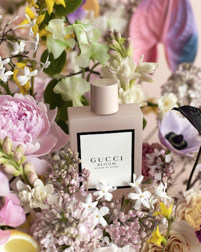 Gucci Bloom perfume bottle in a field of vibrant flowers and blossoms. The light pink bottle stands out amidst the colorful floral arrangement.