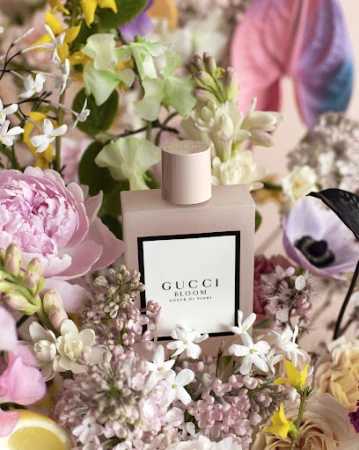 Gucci Bloom perfume bottle in a field of vibrant flowers and blossoms. The light pink bottle stands out amidst the colorful floral arrangement.