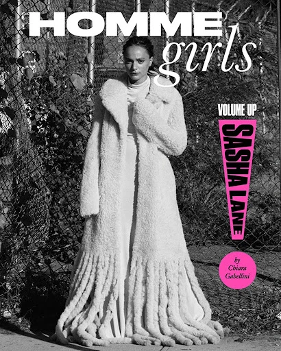 Monochrome fashion magazine cover featuring a woman in a long, fringed coat, against a chain-link fence, with bold text elements.