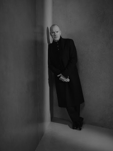 Portrait of a bald adult male in a long black coat leaning against a wall indoors, photographed in black and white.