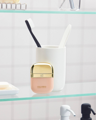 Dolce & Gabbana perfume bottle sits on a glass bathroom shelf next to a toothbrush cup against a white tiled wall, close-up.