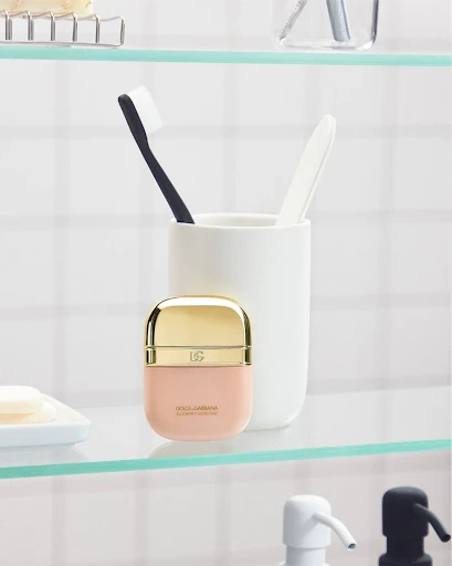 Dolce & Gabbana perfume bottle sits on a glass bathroom shelf next to a toothbrush cup against a white tiled wall, close-up.