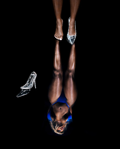 Image of a dark-skinned female model wearing a blue swimsuit, with one foot bare and the other wearing a clear high-heeled shoe.