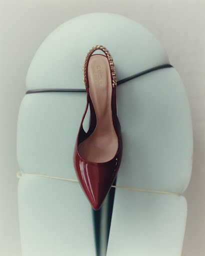 Elegant slingback heel with gold chain strap. Stylish burgundy patent leather shoe rests on an abstract, light blue pedestal for fashion.