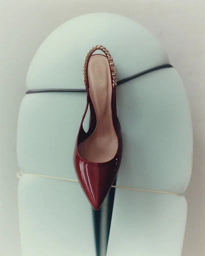 Elegant slingback heel with gold chain strap. Stylish burgundy patent leather shoe rests on an abstract, light blue pedestal for fashion.