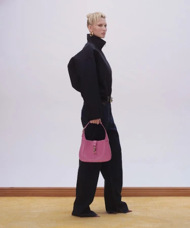 Model in black turtleneck and trousers holding pink handbag against a neutral wall with yellow trim floor