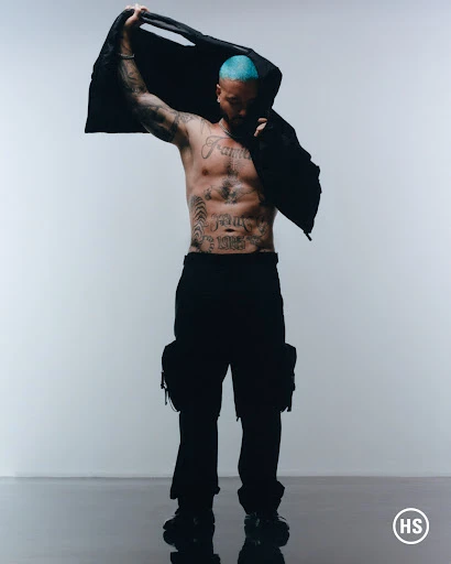 Shirtless male model with blue hair holds jacket, showcasing tattoos against a white background in a studio setting. He is wearing cargo pants.