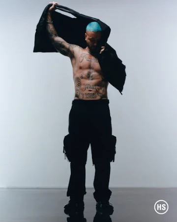 Shirtless male model with blue hair holds jacket, showcasing tattoos against a white background in a studio setting. He is wearing cargo pants.