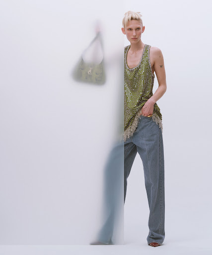 A young adult woman models a green embellished tank top and blue jeans in a studio setting. A blurred figure stands behind her.