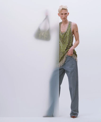 A young adult woman models a green embellished tank top and blue jeans in a studio setting. A blurred figure stands behind her.