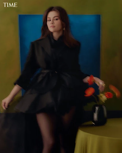 Selena Gomez poses in a black dress and jacket for TIME in front of a teal and yellow paneled background with flowers on a table.