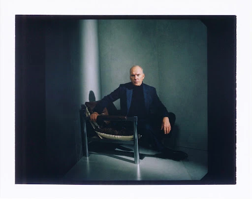 Portrait of a bald adult male in a blazer sitting in a metal chair in a photo studio with a grey background and a light shaft.