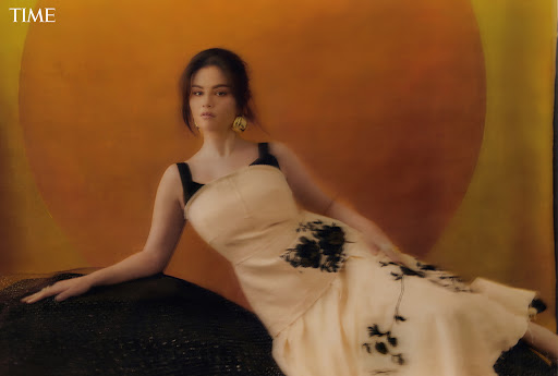 Woman in a light dress reclines against a black textured object in a warm-toned photo studio, with a large orange circle backdrop.