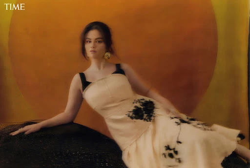 Woman in a light dress reclines against a black textured object in a warm-toned photo studio, with a large orange circle backdrop.