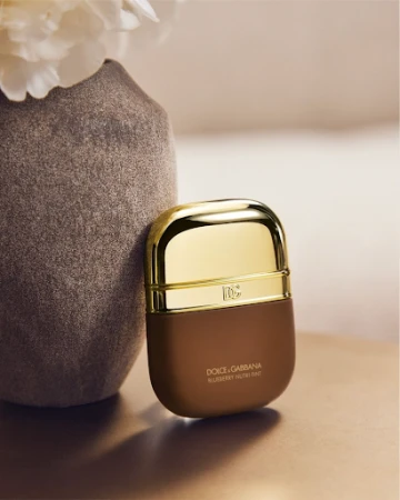 Dolce & Gabbana compact makeup case in brown and gold resting against a textured grey ceramic vase with white flowers.