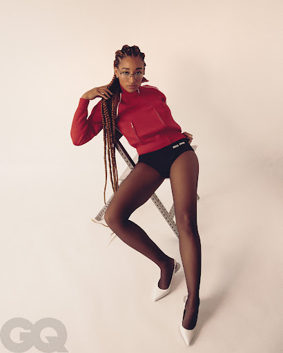Woman with cornrows in red sweatshirt, black briefs and sheer tights sitting on a metal stool in a white photo studio.