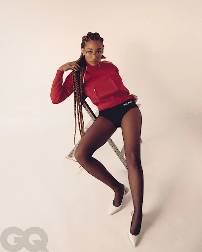 Woman with cornrows in red sweatshirt, black briefs and sheer tights sitting on a metal stool in a white photo studio.