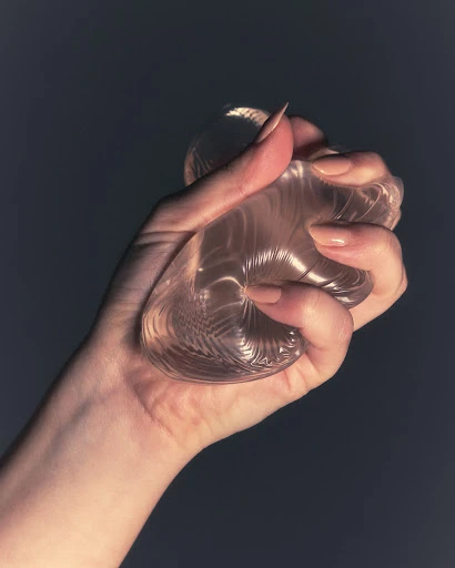 A hand squeezes a clear silicone object against a dark background, the long fingernails are manicured with neutral colored polish.