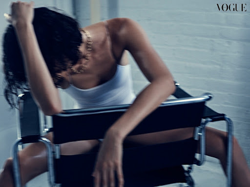 A fashionable adult female model poses while seated in a chair, bathed in studio lighting, highlighting her tank top and gold necklace.