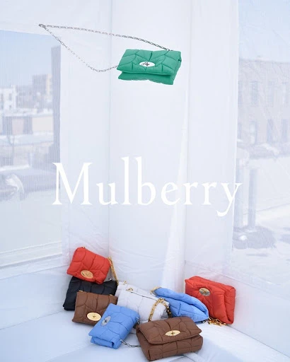 Collection of colorful quilted purses with chain straps, in an indoor setting with white curtains, and natural light, logo present.