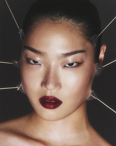 Portrait of an Asian woman with dark red lipstick and wooden sticks taped to her face against a dark background in a beauty shot.