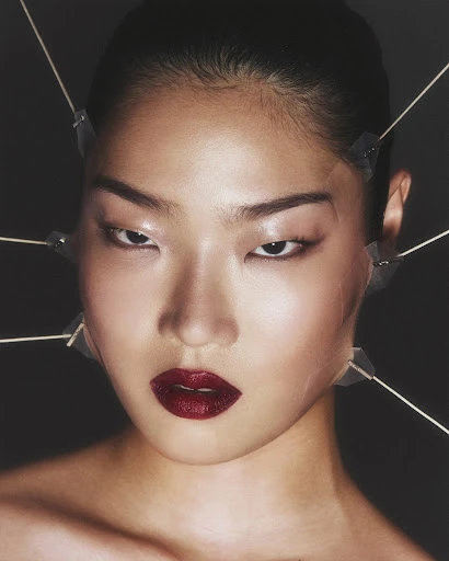 Portrait of an Asian woman with dark red lipstick and wooden sticks taped to her face against a dark background in a beauty shot.