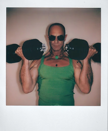Polaroid photo: man in tank top lifts dumbbells indoors. He wears sunglasses and a necklace, set against a plain background.