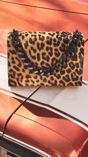 Spotted handbag with black chain detail sits on the hood of a shiny orange car, highlighting texture and color.