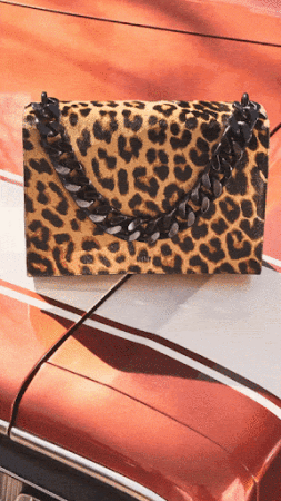 Spotted handbag with black chain detail sits on the hood of a shiny orange car, highlighting texture and color.