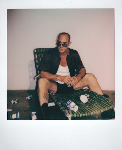 Man in sunglasses relaxes in a lawn chair surrounded by discarded cups in an indoor setting with a neutral background.