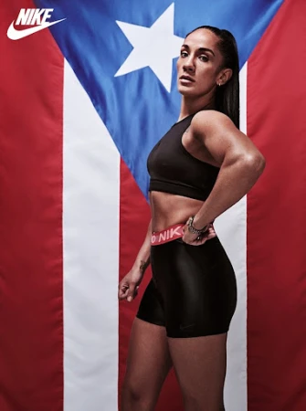 Fit woman in workout clothes poses in front of a Puerto Rican flag indoors. Nike logo visible in the upper left corner.