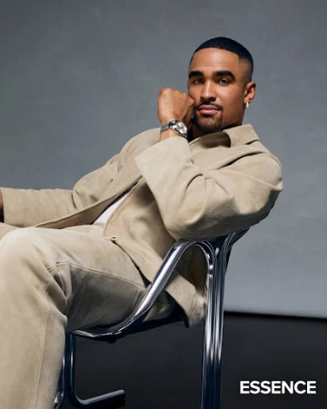 Jalen Hurts seated in a chrome-framed chair wearing a beige jacket and trousers against a grey textured studio backdrop.