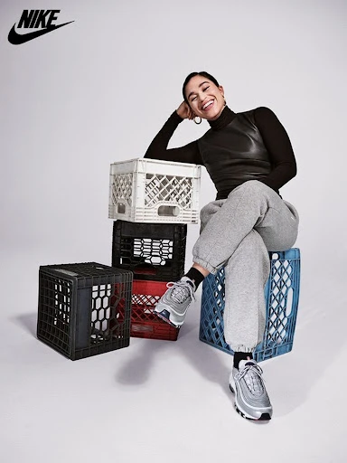 Smiling woman sits on milk crates wearing grey sweatpants, black turtleneck top, and silver Nike sneakers in a studio shot.