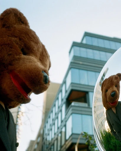 Urban shot of a person wearing a teddy bear head mask and a suit, reflected in a metallic spherical object, against a city backdrop.