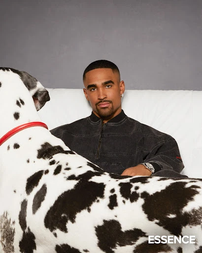 Portrait of an adult male sitting on a white couch next to a Great Dane dog in a photo studio against a solid grey backdrop.