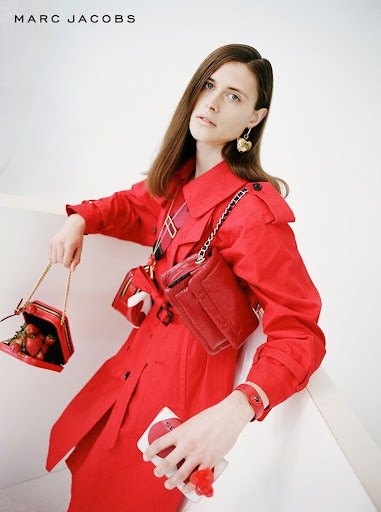 Fashion shot of a woman in a red trench coat, holding strawberry purse and a compact, posing against a bright white background.
