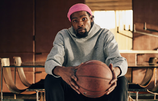 Portrait of an adult Black male in a pink beanie and grey hoodie, holding a basketball, in a naturally lit indoor environment.