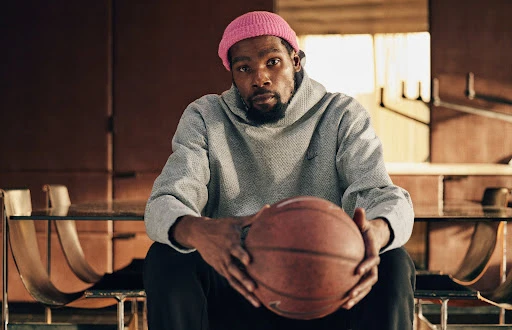 Portrait of an adult Black male in a pink beanie and grey hoodie, holding a basketball, in a naturally lit indoor environment.