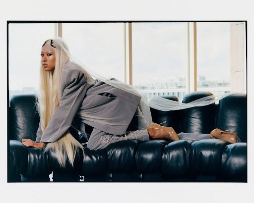 Fashionable woman in grey suit and sheer veil poses on black leather couch in a light-filled room with city views. Stylish studio portrait.