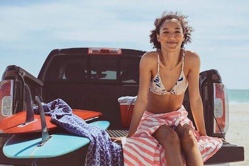 Smiling woman in bikini sits on pickup truck bed on sunny beach with surfboards and towels. Travel, summer, lifestyle scene.