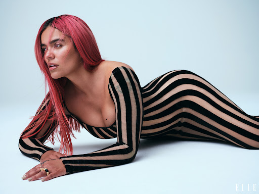 Latin woman Karol G posing in a photo studio wearing a striped body suit while laying down on a light blue surface.